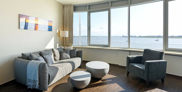 Bright hotel room with gray sofa, armchairs, round tables and panoramic window overlooking the water.
