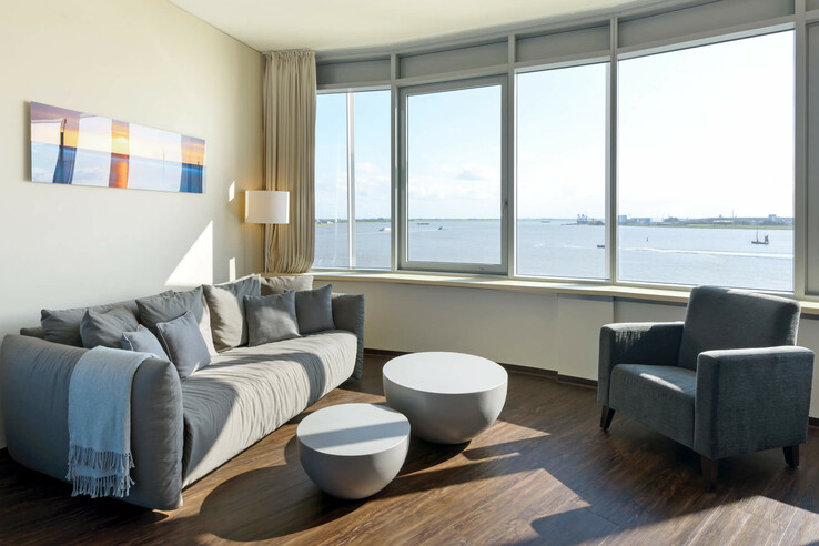 Bright hotel room with gray sofa, armchairs, round tables and panoramic window overlooking the water.