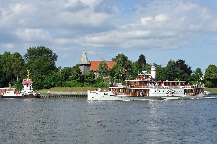 Dampfschiff auf der Trave mit grüner Uferlandschaft und Kirche im Hintergrund bei sonnigem Wetter.