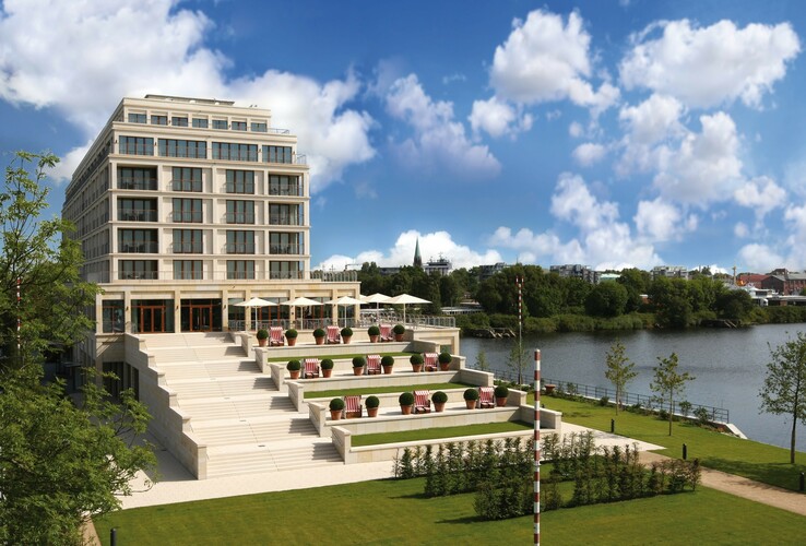 Modernes Hotel am Fluss mit großen Fenstern, gestuften Terrassen und Liegestühlen, umgeben von grüner Landschaft.