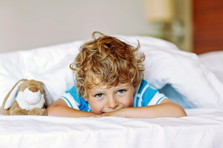 Kind mit lockigem Haar liegt lächelnd neben Kuscheltier auf weißem Bett in einem Hotelzimmer.