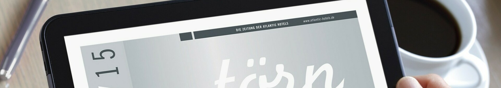 Tablet mit Hotelmagazin "törn" auf dem Bildschirm, gehalten von Händen, daneben eine Tasse Kaffee auf Holztisch.