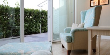 Comfortable armchair with cushions in bright room, view of terrace with green hedge through glass door.