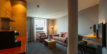 Modernes Hotelzimmer mit Sofa, Sessel, Schreibtisch, Fernseher und Fenster; warme Beleuchtung und dezente Farben.