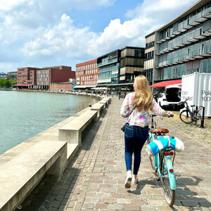 Woman pushing bicycle along the water, modern buildings in the background, in sunny weather.