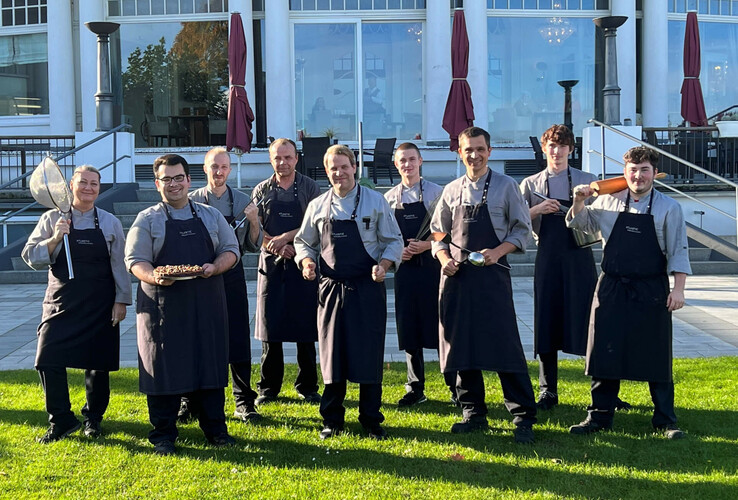 The kitchen team of the ATLANTIC Grand Hotel Travemünde poses smiling in uniforms in front of the hotel building.
