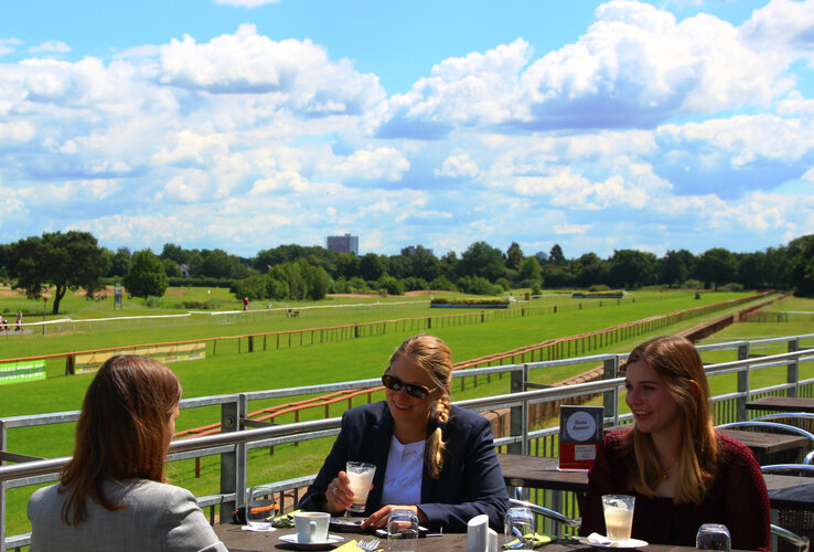 Sonnenterrasse des Restaurant DERBY Drei Personen genießen Getränke auf einer Terrasse mit Blick auf die grüne Galopprennbahn bei sonnigem Wetter.