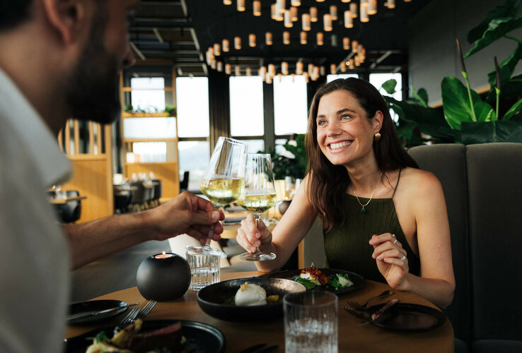 Paar stößt im eleganten Restaurant des ATLANTIC Hotel Heidelberg mit Weißwein an, umgeben von stilvoller Dekoration.
