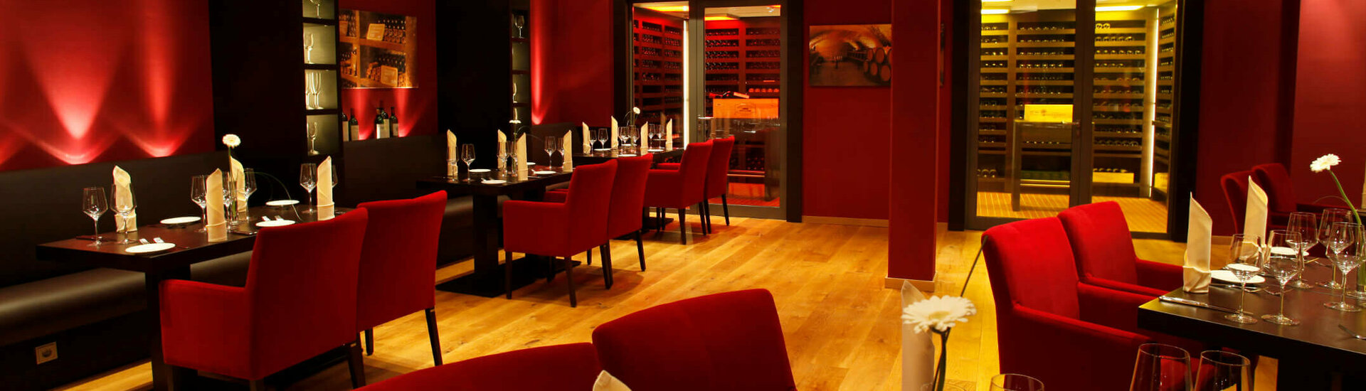 Elegant restaurant with red walls and chairs, stylishly laid tables and an illuminated wine cabinet in the background.