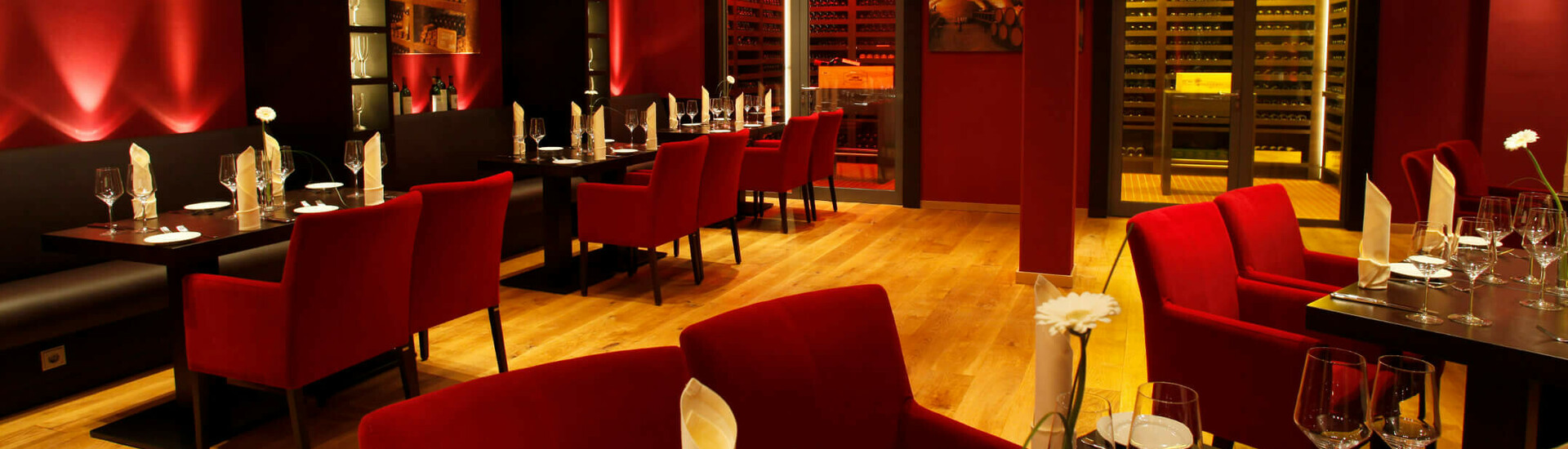 Elegantes Restaurant mit roten Wänden und Stühlen, stilvoll gedeckten Tischen und beleuchtetem Weinschrank im Hintergrund.