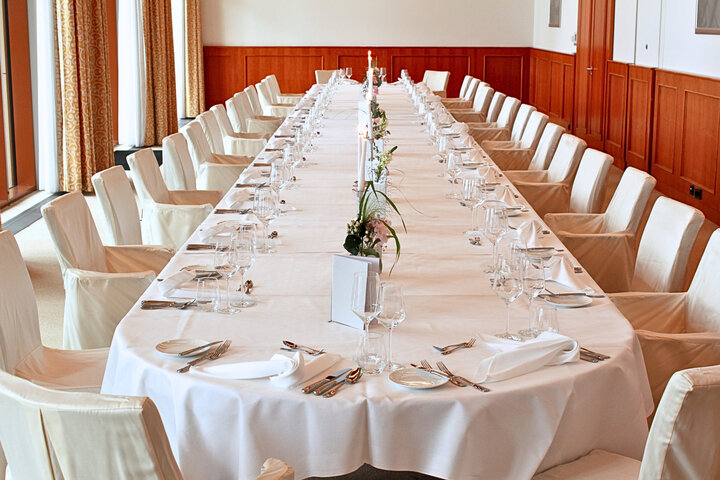 An oblong table laid for a festive occasion, with a white tablecloth, wine glasses, napkins, cutlery and chairs with fabric covers.